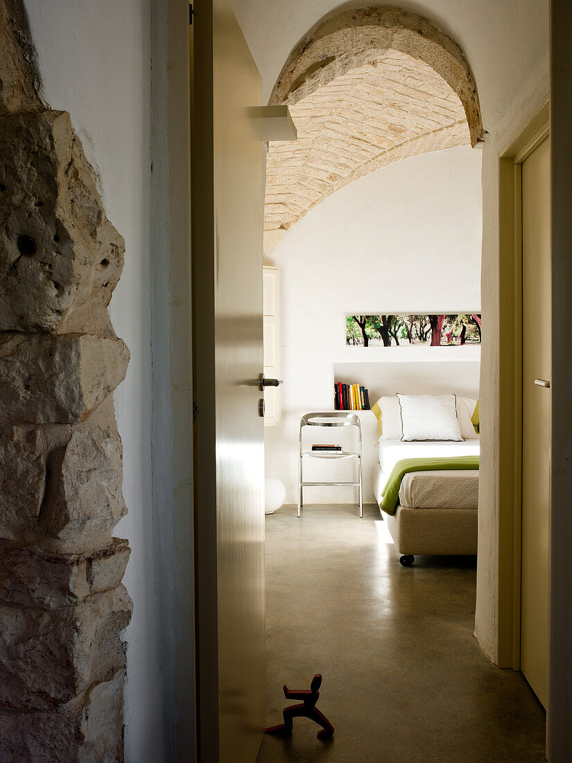 View through open anteroom door of metal chair next to modern double bed in bedroom with brick vaulted ceiling