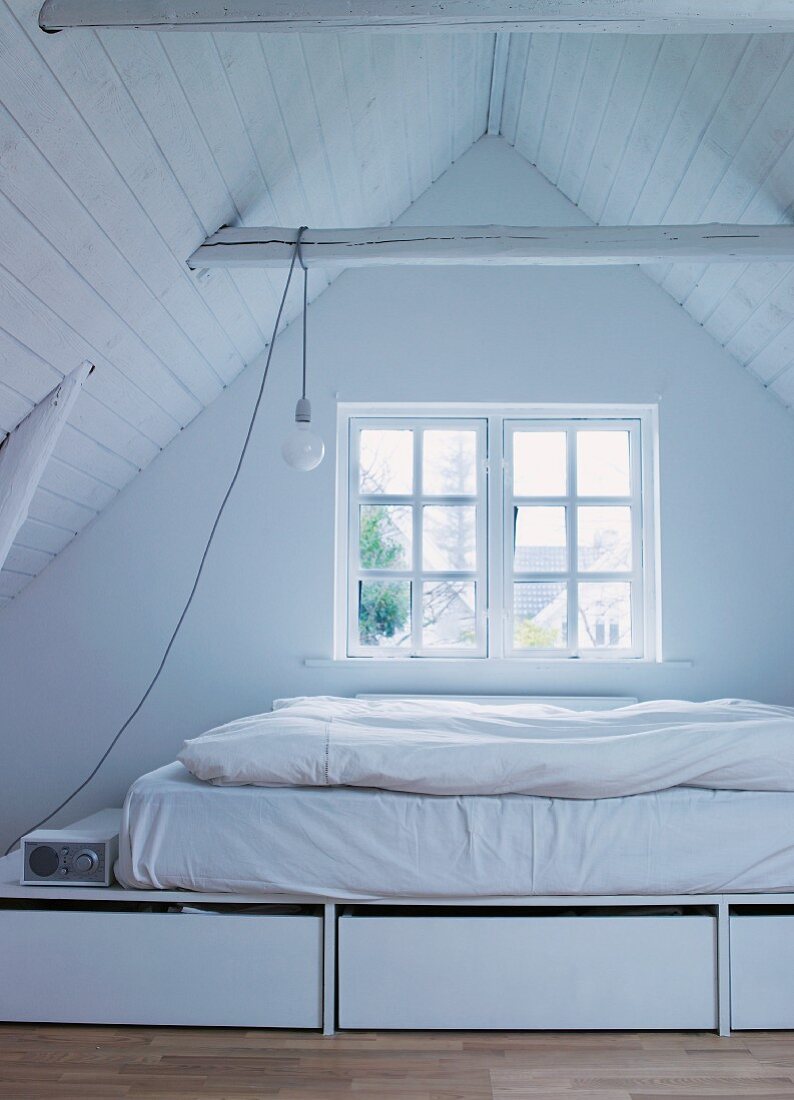 Attic room with mattress and bedding on white platform with drawers in front of window