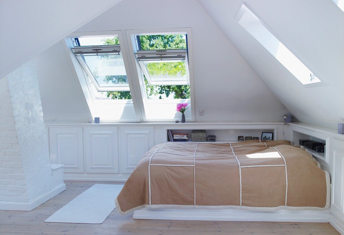Bett mit Tagesdecke in heller Dachzimmerecke und offen stehenden Dachfenstern
