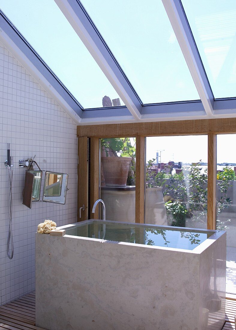 Cubist bathtub full of water below sloping glass roof and with view through terrace window