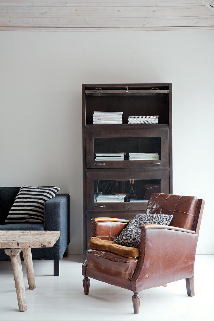Worn leather armchair in front of vintage office cabinet with fold-down glass doors and rustic wooden table in front of modern sofa