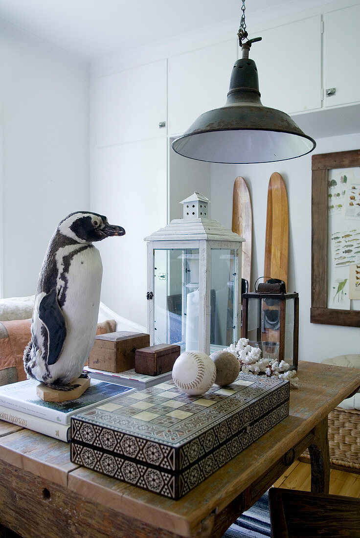 Ausgestopfter Pinguin auf Bücherstapel, weiße Laternenleuchte und Brettspiel auf rustikalem Holztisch unter Vintage Hängeleuchte