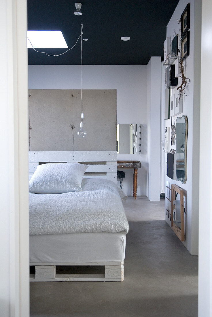 Blick in ein Loft-Schlafzimmer mit schwarzer Decke - ein Selbstbaubett aus weiss gestrichenen Europaletten vor einer Trennwand aus Sichtbeton