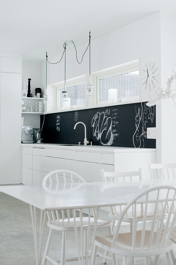 Offene Designerküche in puristischem Weiß mit schwarzem Spritzschutz und weiss lackierten Designerstühlen am Esstisch