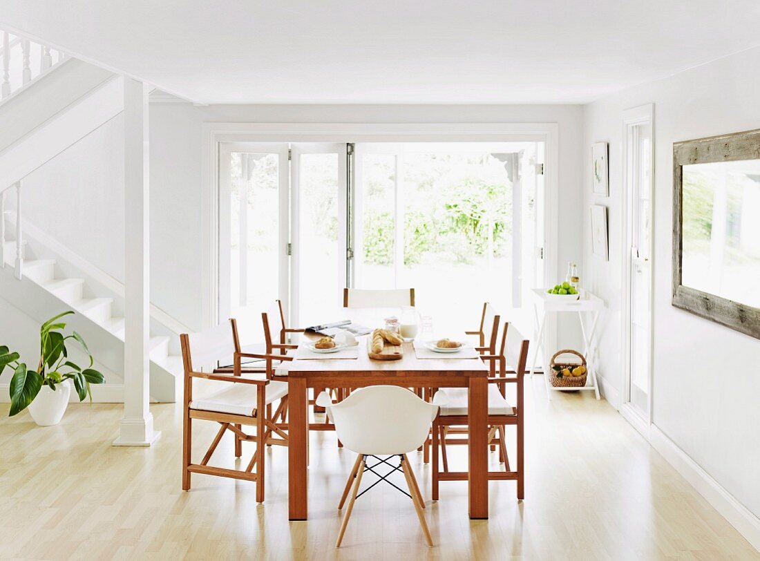 Wooden dining table and matching chairs in front of terrace windows in open-plan, white interior with staircase