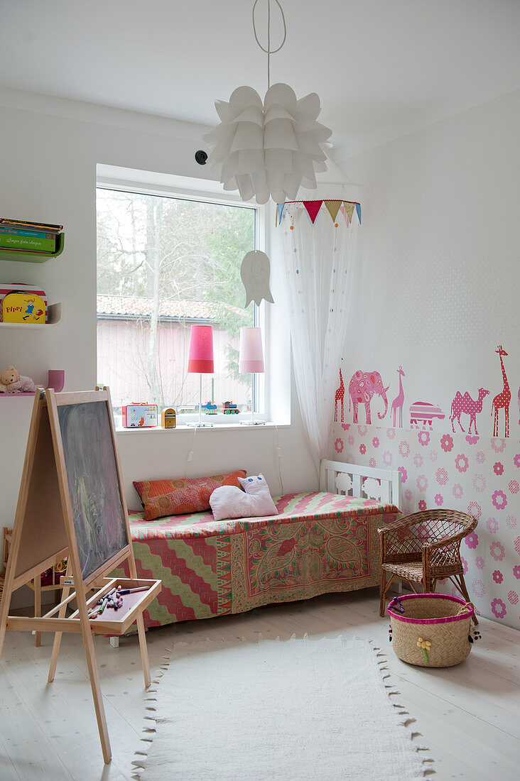 Aufgestellte Schreibtafel vor dem Bett mit gemusterter Tagesdecke am Fenster und bemalter Wand in weißem Kinderzimmer