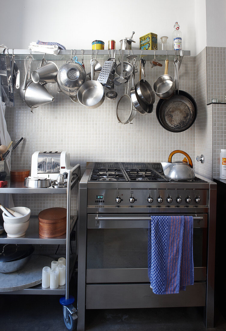 Moderner Gasherd mit Edelstahlfront neben Rolltisch und Wandbord mit aufgehängtem Kochgeschirr