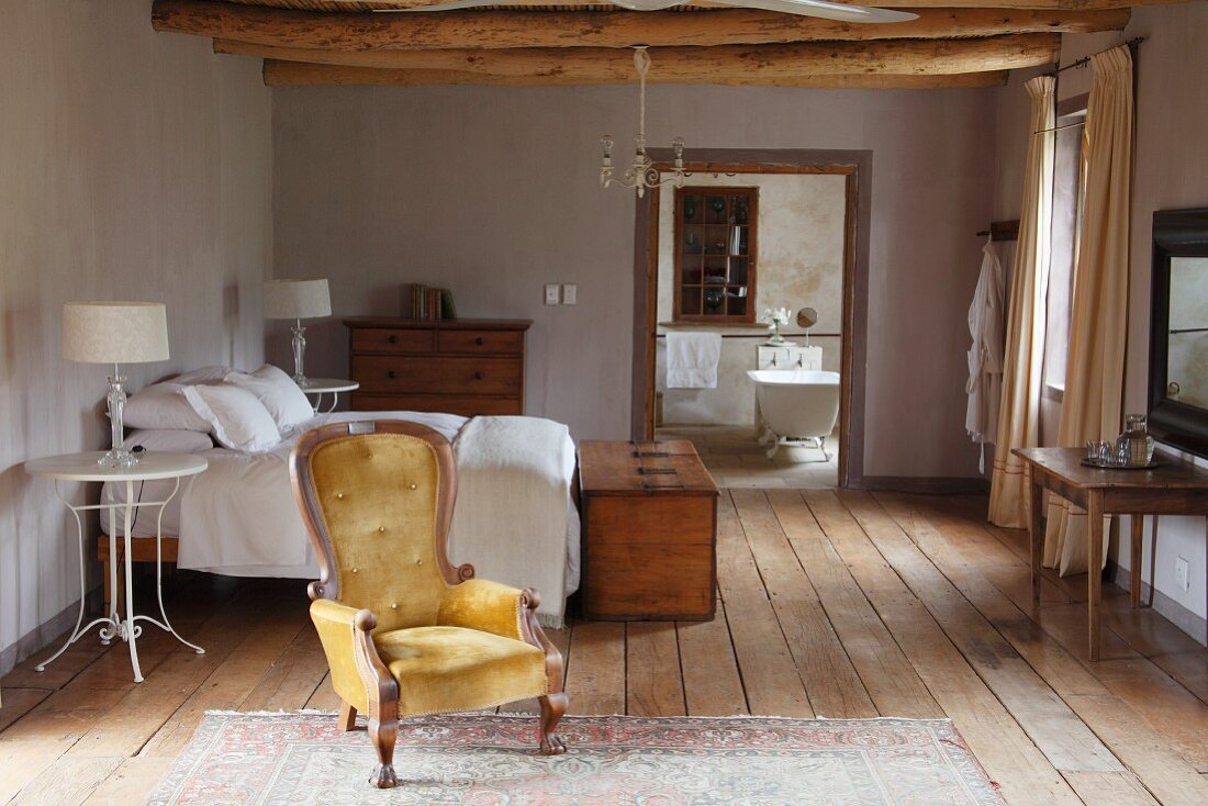 Antiker Sessel mit goldgelbem Bezug vor Doppelbett in rustikalem Ambiente und Blick in offenes Bad