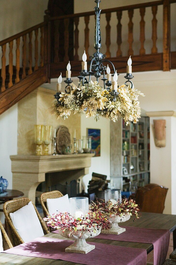 Decorated metal chandelier above dining table with flower arrangements in bowls in open-plan foyer with staircase and gallery
