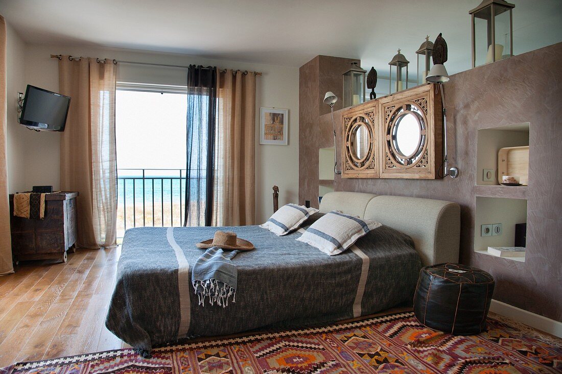 Doppelbett vor gemauertem Raumteiler mit aufgehängtem Spiegel und offene Balkontür mit Meerblick