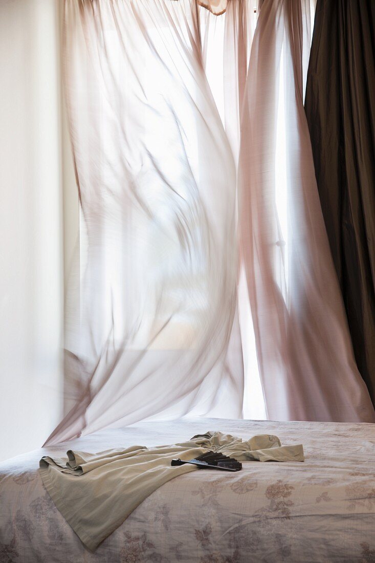 Bett vor verhülltem Fenster mit wehendem, luftigem Vorhang