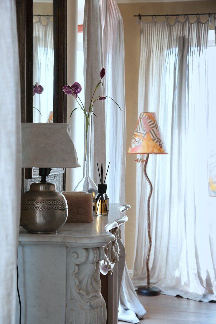 Tischlampe auf Kaminsims eines offenen herrschaftlichen Kamins und moderne Stehleuchte in Wohnzimmerecke neben Fenster mit luftigem Vorhang