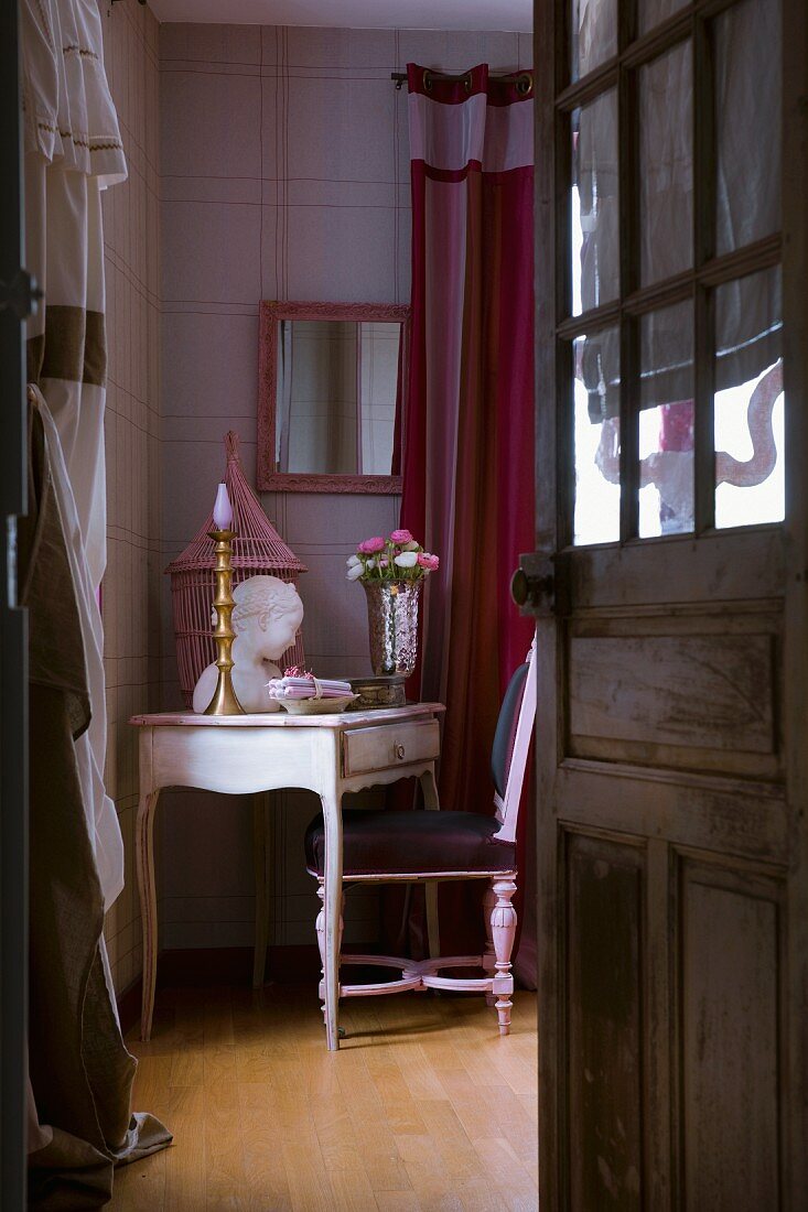 Blick durch eine geöffnete Holztür mit Glasfüllung auf zierlichen antiken Schreibtisch mit femininer Dekoration in Rosetönen