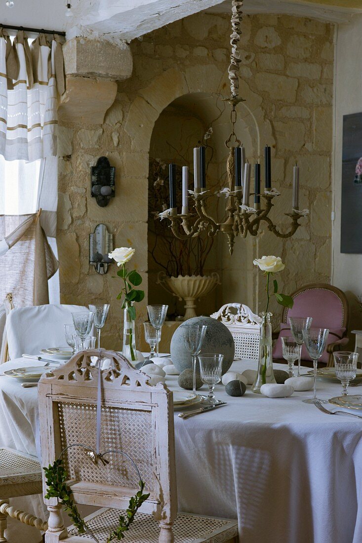 Messing-Kerzenleuchter über festlich gedeckter Tafel mit antiken, weissen Stühlen im Vintage-Look; Natursteinwand im Hintergrund