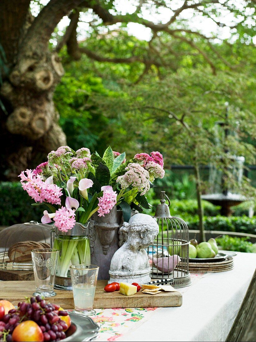 Büste, Steinvase mit Blumen, Vogelkäfig und Obstteller auf einem Tisch im Garten