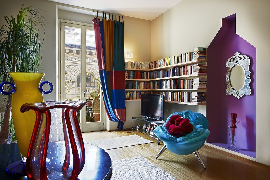 Modernes Wohnzimmer mit postmodernen Akzenten - blauer Plüschsessel vor violetter Wandnische und farbige Glasvasen auf Tisch