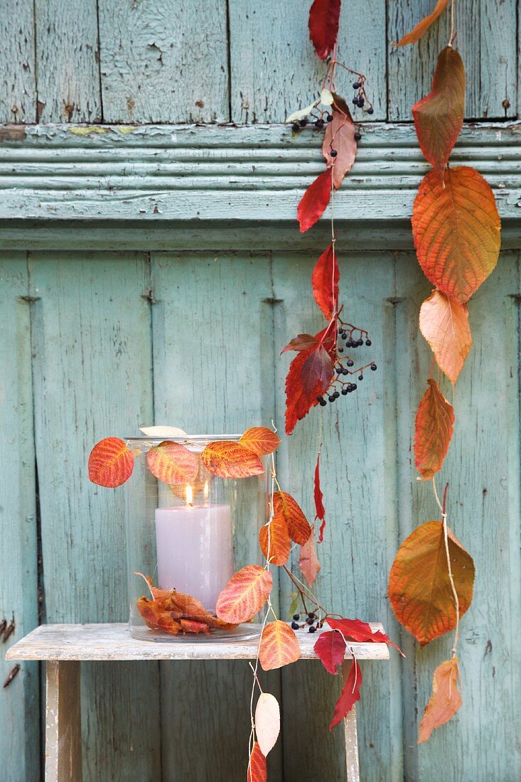 Aufgefädelte Herbstblätter und Windlicht vor Holzwand