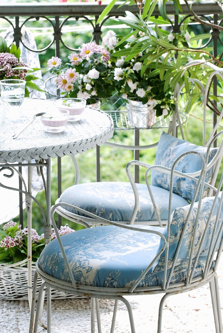 weiße Metallstühle mit Polsterkissen und Bistrotisch vor schmiedeeisernem Geländer mit Blumen in Hängegitter