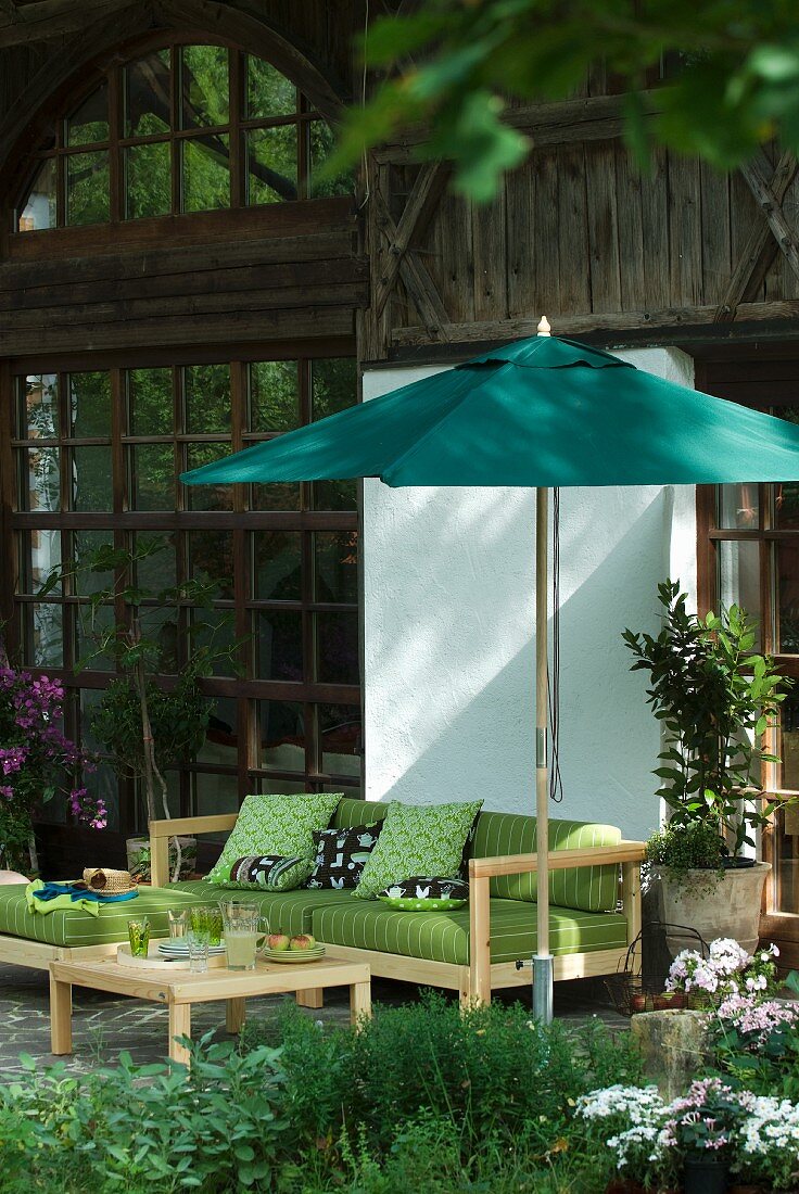 Gemütliches Terrassensofa mit grünem Polster unter Sonnenschirm vor rustikalem Bauernhaus