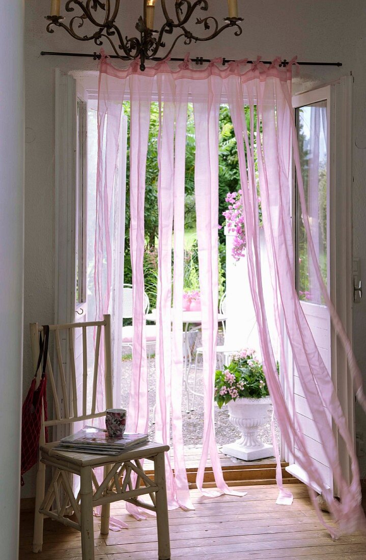 Blick auf weiss lackierten Bambusstuhl und schmiedeeiserenen Kronleuchter vor wehendem, romantischem Bahnen-Vorhang an der offenen Terrassentür
