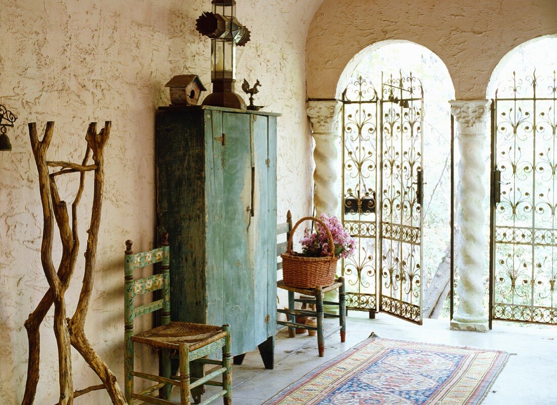 Antiker Holzschrank zwischen traditionellen Bauernstühlen in einem Arkadengang mit ornamentiertem Eisenzaun