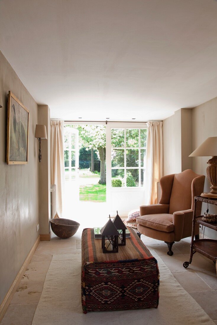 Schmaler Raum mit offener Tür zum Garten; traditioneller Ohrenbackensessel und Windlichter auf Polstertisch mit Kelimmuster
