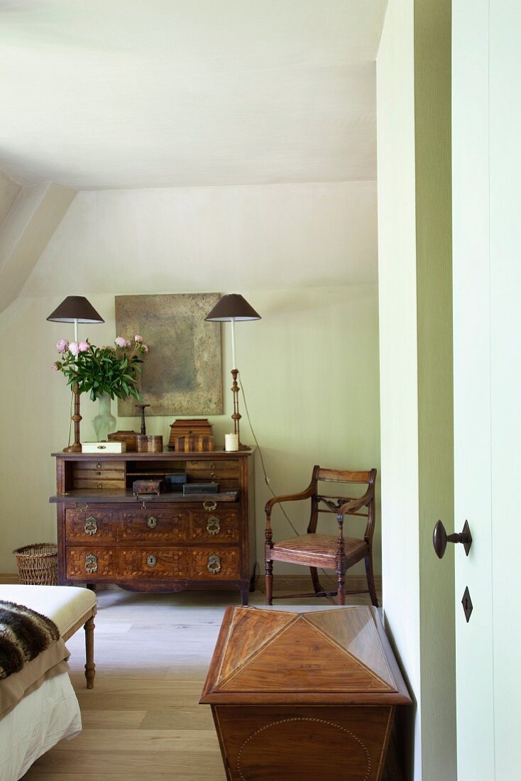 Comfortable attic bedroom with antique furniture - armchair next to unusual, tall table lamps on bureau below modern painting