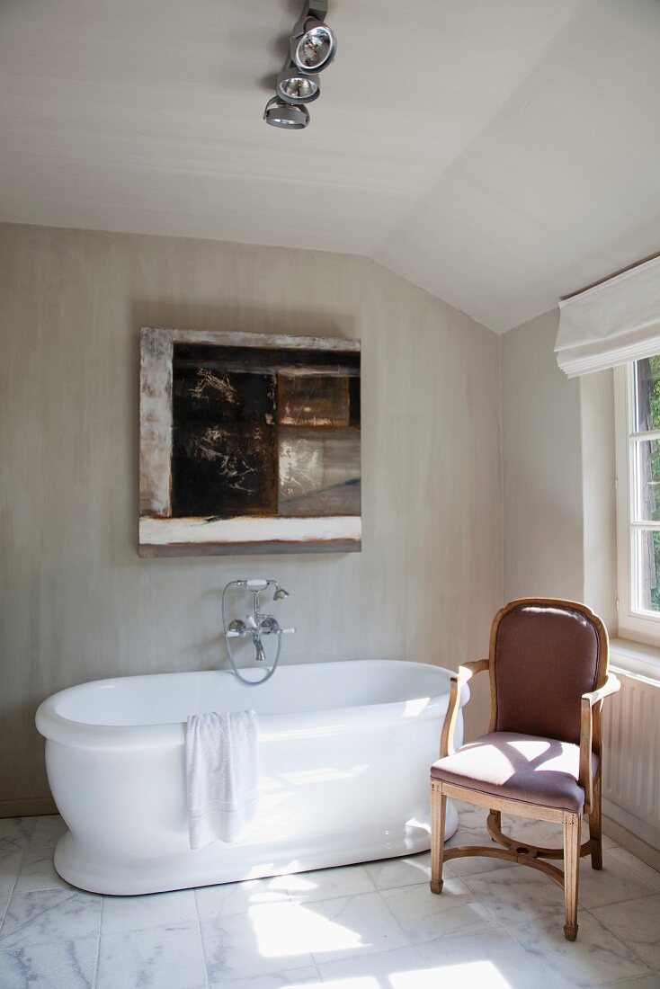 Bathroom with attic ceiling and veined marble floor; modern painting over free-standing bathtub next to antique upholstered chair