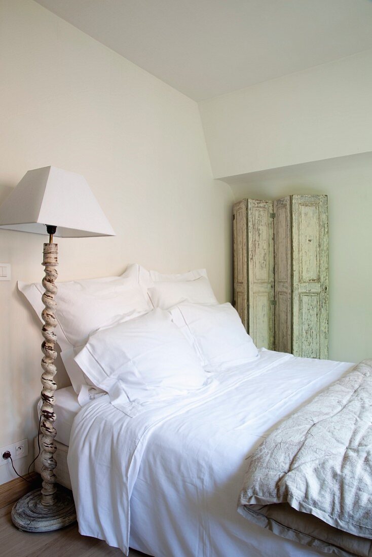 Einfaches Mansardenzimmer mit klassisch weiss bezogenem Bett, abgeblättertem Paravent an der Wand und Stehlampe im antiken Vintagelook