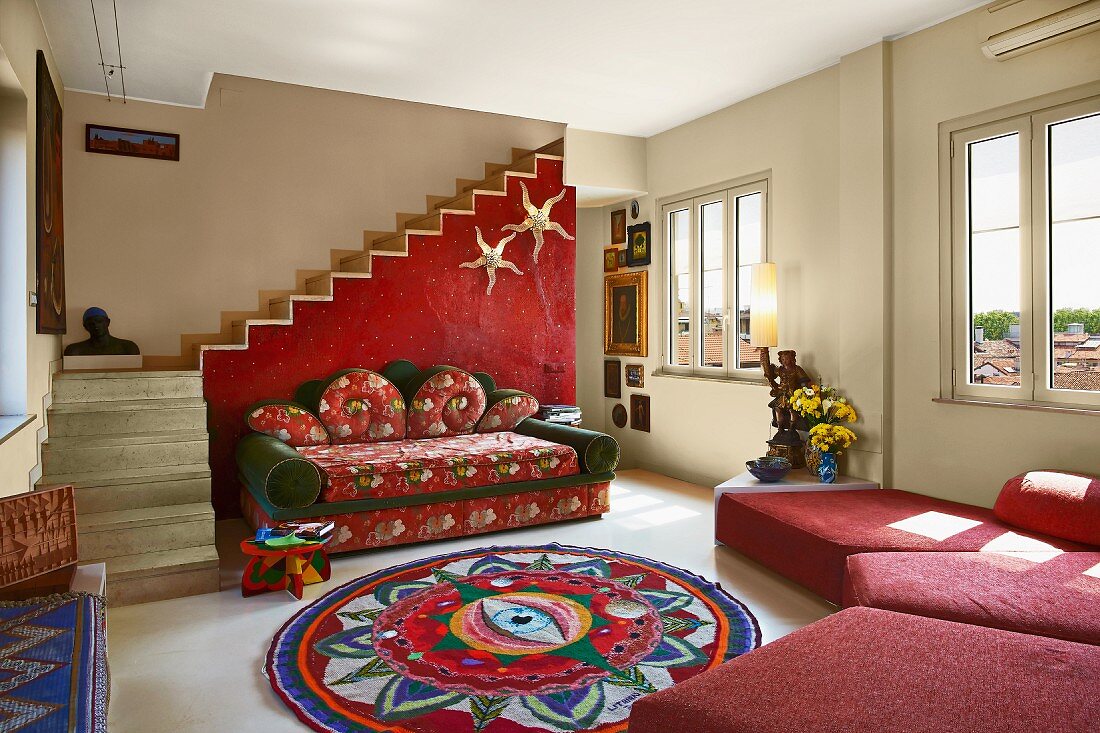 Gemütliche Sofalandschaft in Rot und bunter Teppich im Ethnolook in schlichtem Wohnraum mit Treppe