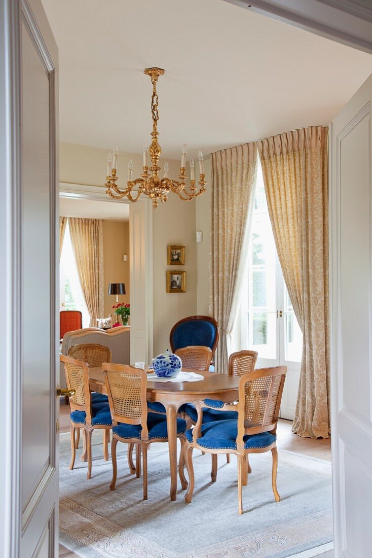 View through open door into dining room with Rococo chairs with blue seats in front of French windows with gathered curtains