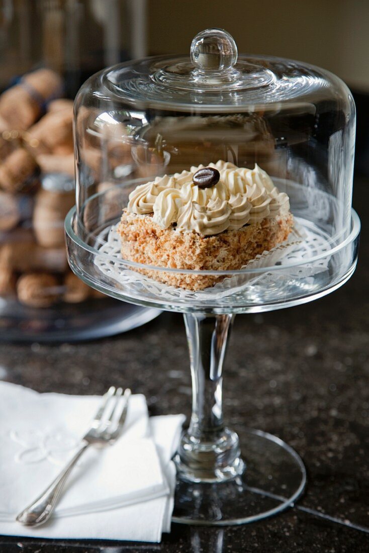 A portion of cake topped with cream and a coffee bean, under a glass dome, on a glass stand with a stem