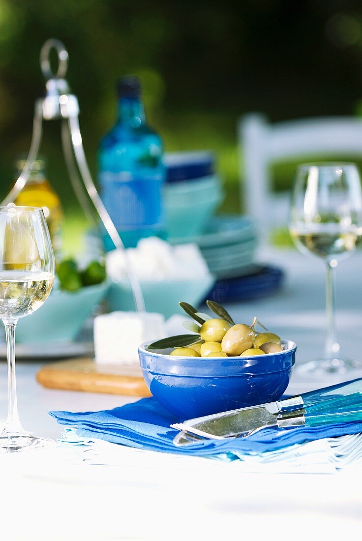 Griechische Oliven in blauem Schälchen, Weingläser und Schafskäse auf gedecktem Tisch