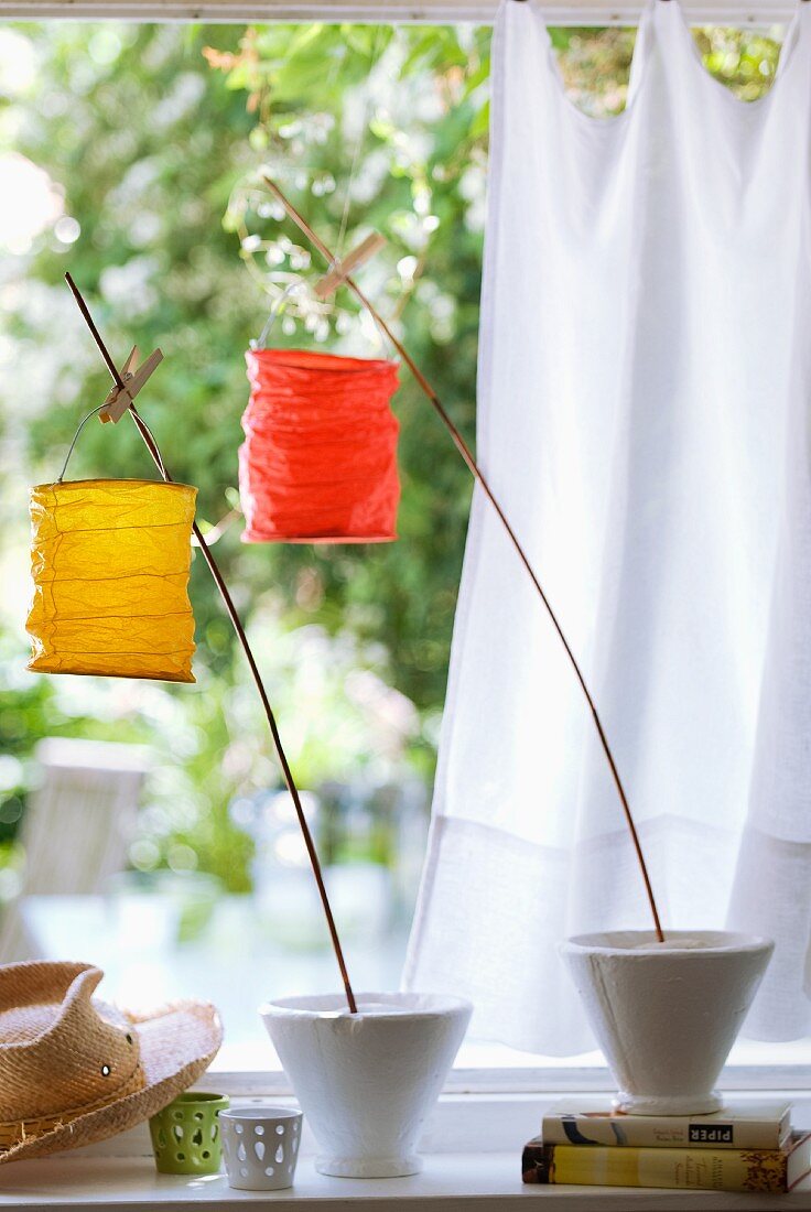 Kleine Papierlampions, mit Wäscheklammern an Holzstäben befestigt, in Gartenatmosphäre mit weißem Vorhang im Hintergrund