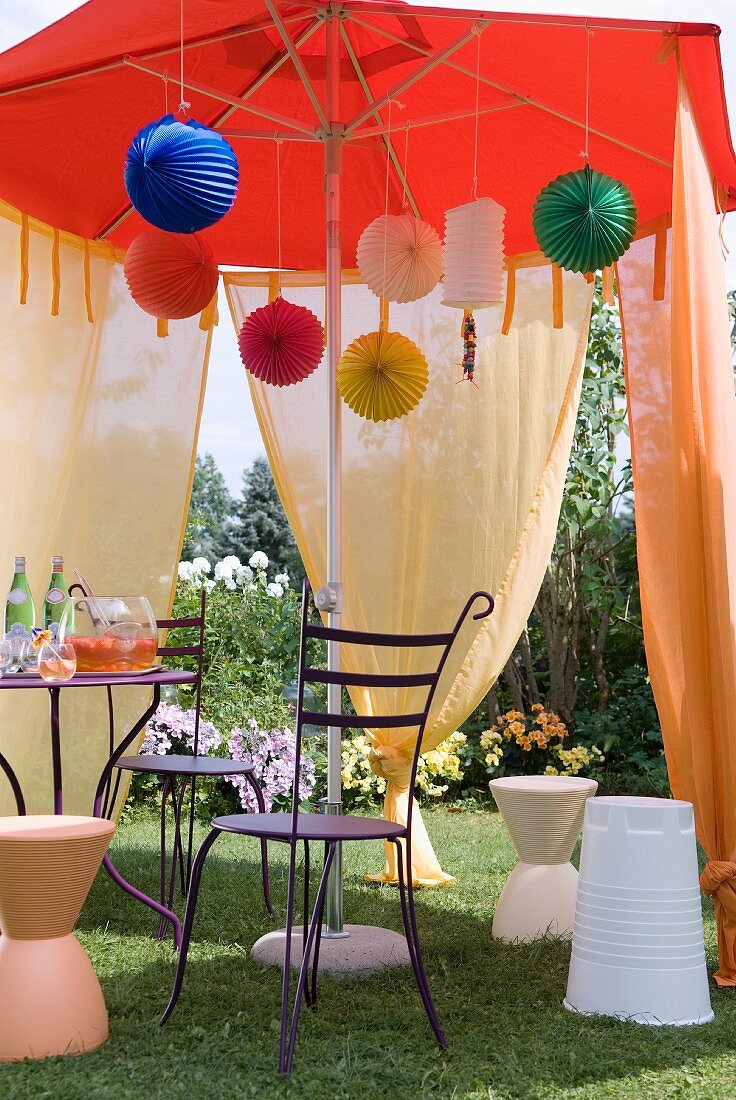 Bistro table with metal chairs and colourful paper lanterns below market umbrella draped with voile curtains