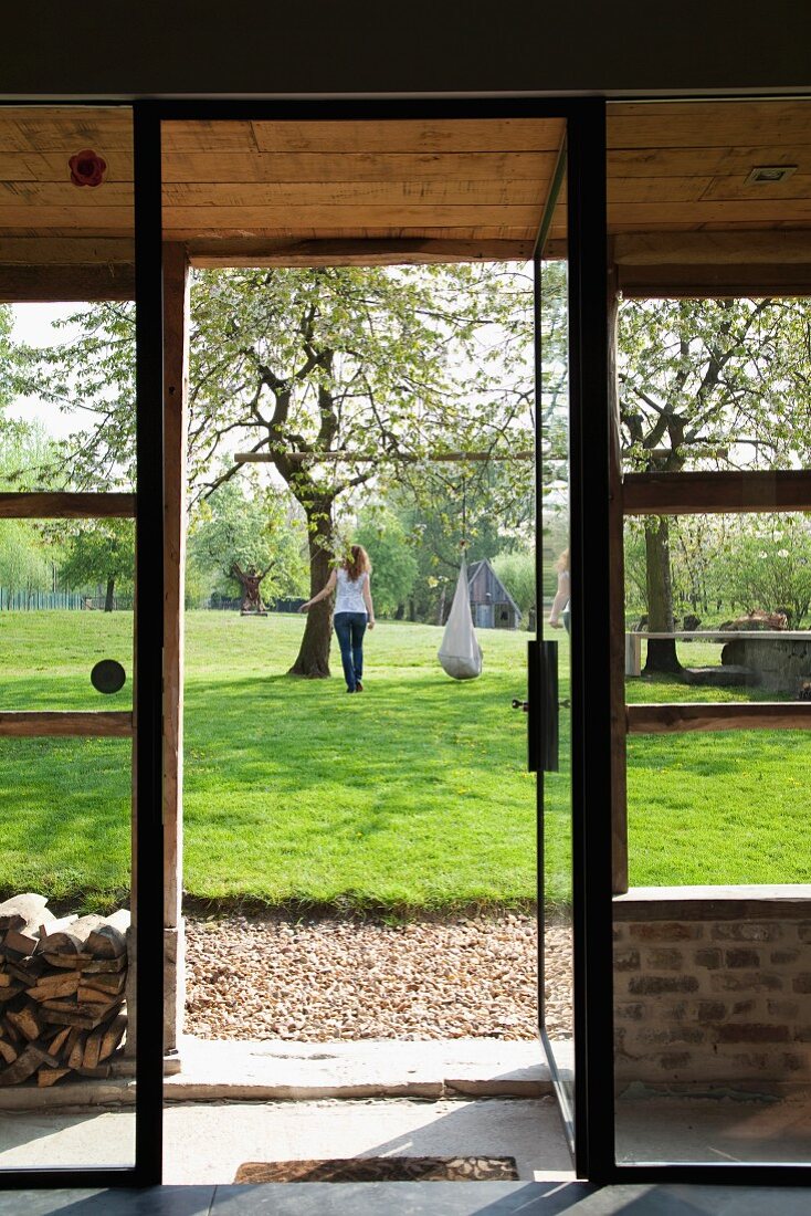 Blick durch offene Glastür eines modernen Wohnhauses auf Frau im frühlingshaften Garten