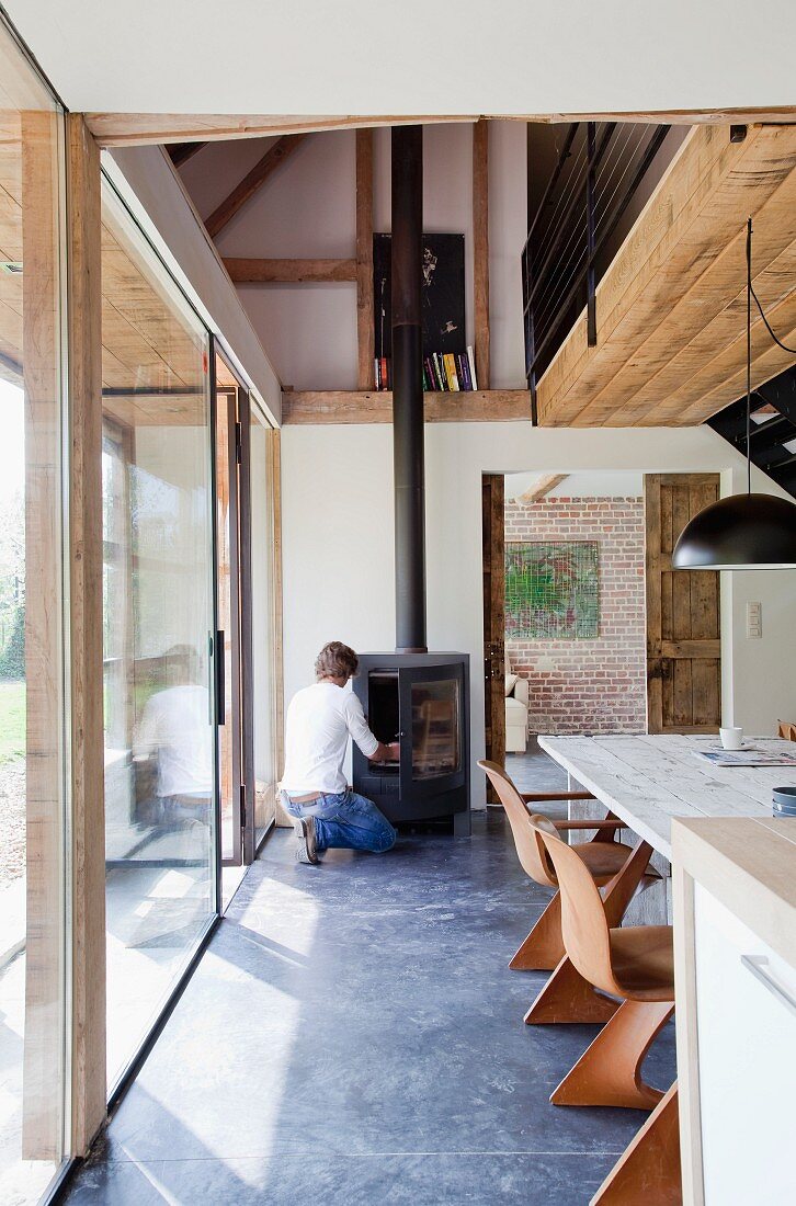 Wooden shell chairs at dining table in modern dining room with view of gallery; man stoking wood burner