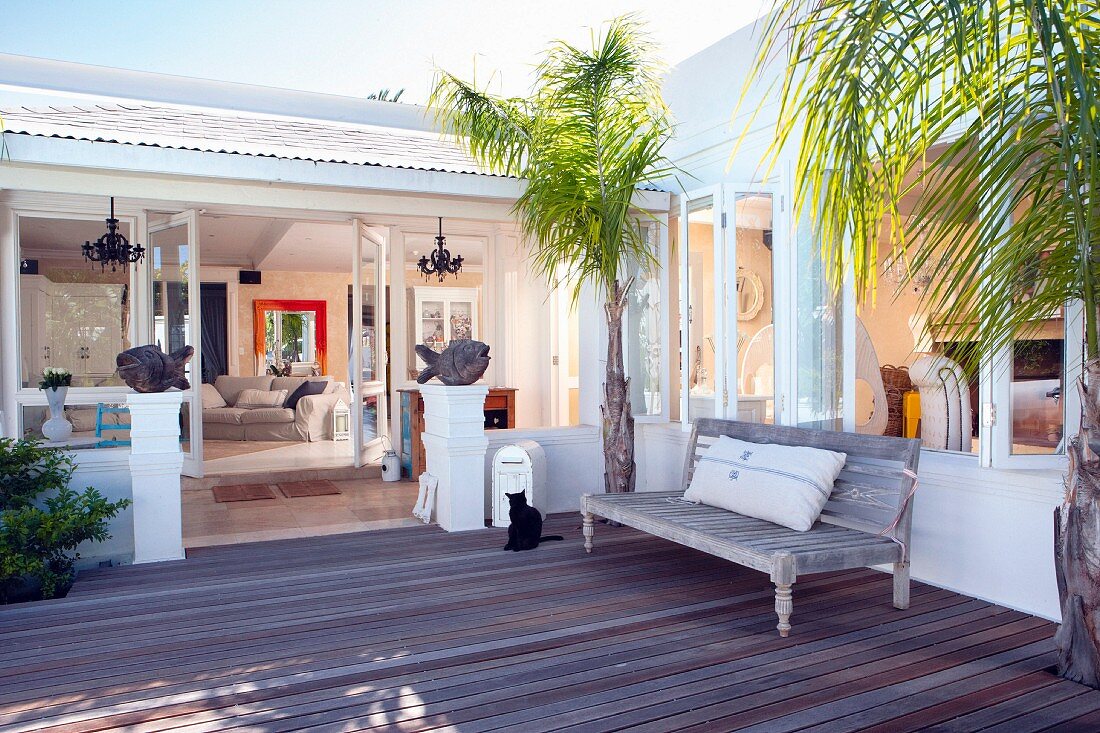 Sitzbank aus Holz zwischen Palmen auf moderner Holzterrasse vor mediterranem Haus mit offenen Terrassentüren
