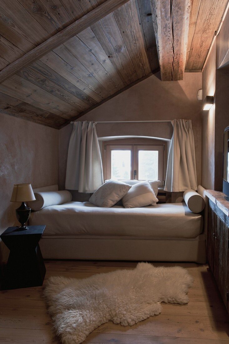 Einfaches, gemütliches Schlafzimmer unter der Dachschräge mit kleinem Fenster; am Boden ein weicher Fellteppich