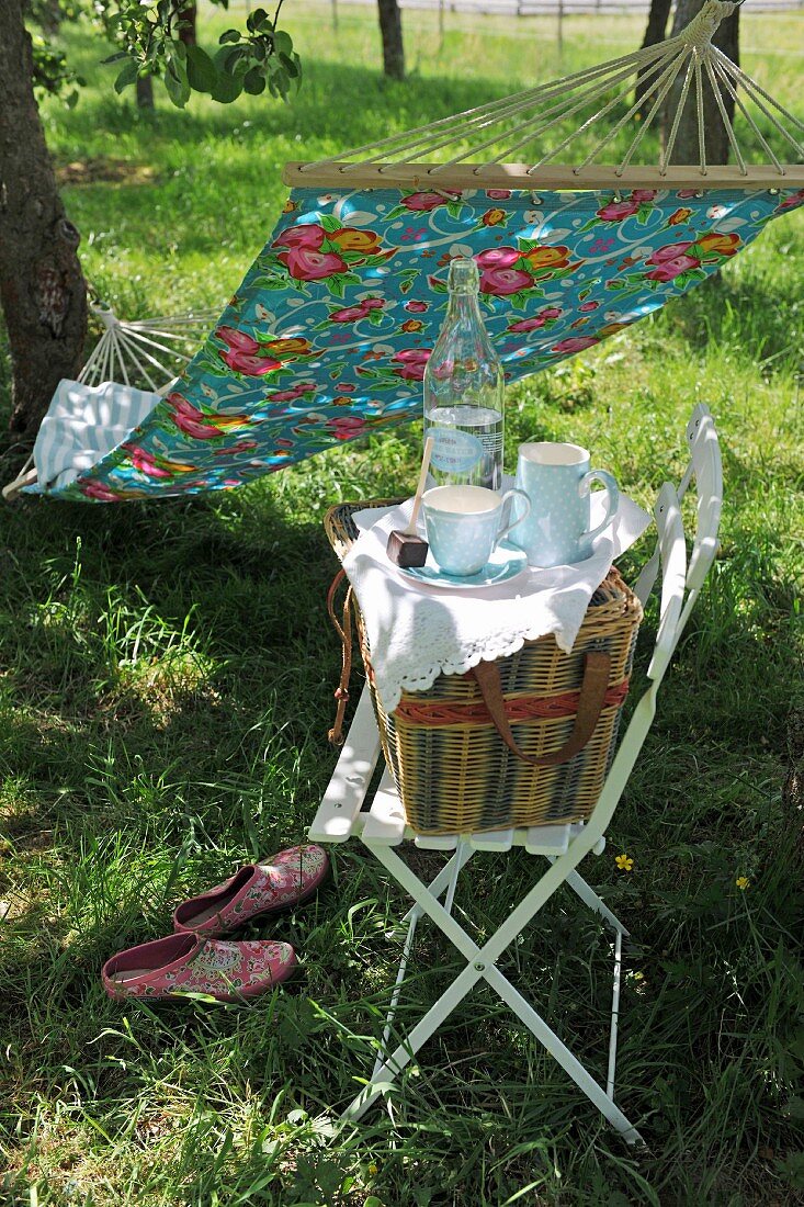 Geschirr und Picknickkorb auf Stuhl vor aufgehängter Hängematte im Garten
