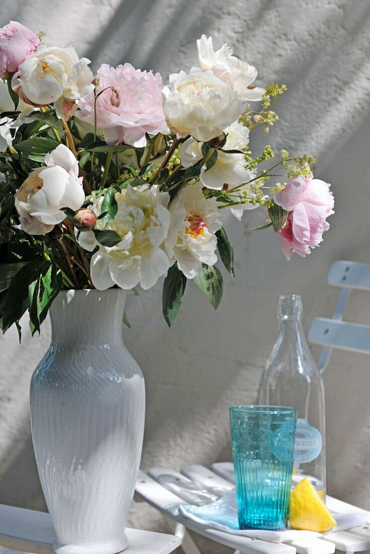 Sommerblumenstrauss in weisser Porzellanvase neben Gartenstuhl mit Glas und Flasche im Retrostil