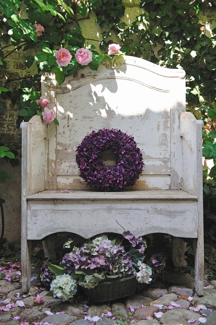 Verwitterte Gartenbank mit Blumenkranz aus violetten Blüten und Blumenschale auf Steinboden