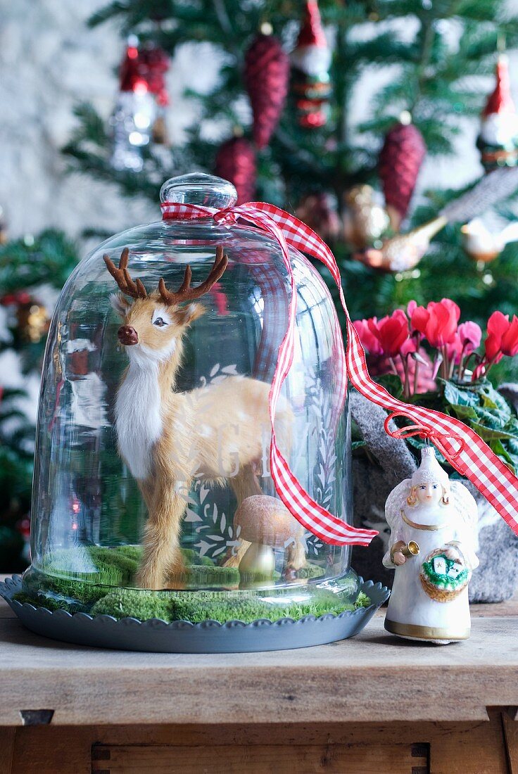 Weihnachtsdeko - Tierfigur unter Glashaube und Engelfigur auf Holzablage vor geschmücktem Weihnachtsbaum