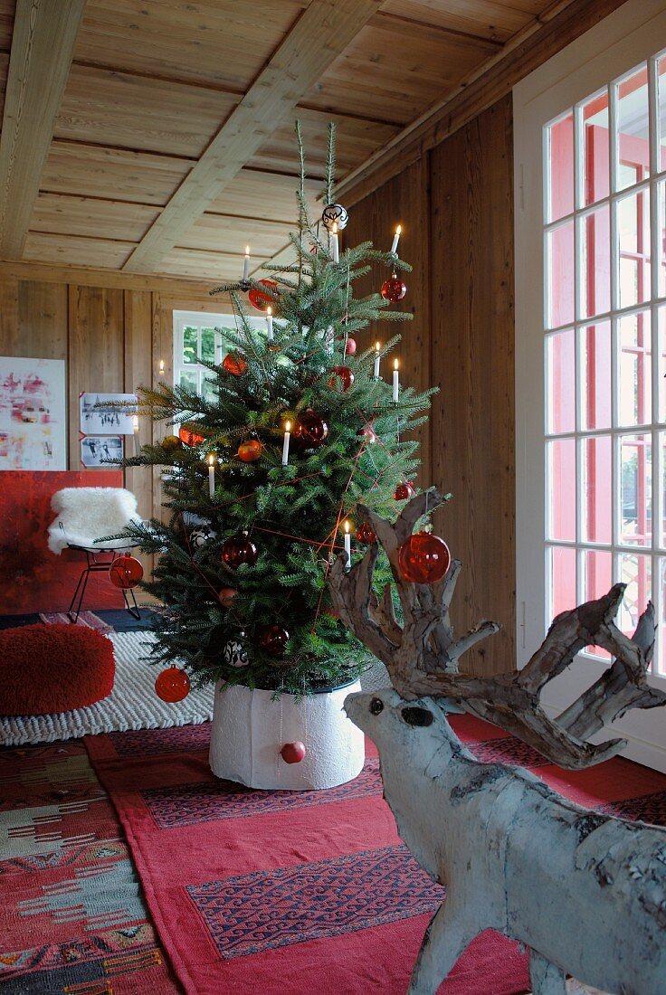 Weihnachtsbaum mit roten Glaskugeln und Kerzen geschmückt in einem Zimmer mit Holzwänden