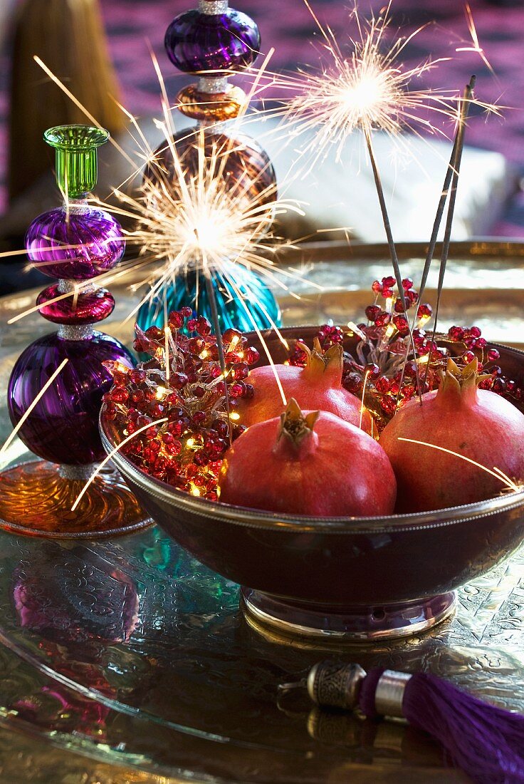 Granatäpfel mit brennenden Wunderkerzen in einer Schale und Kerzenständer aus buntem Glas im Hintergrund