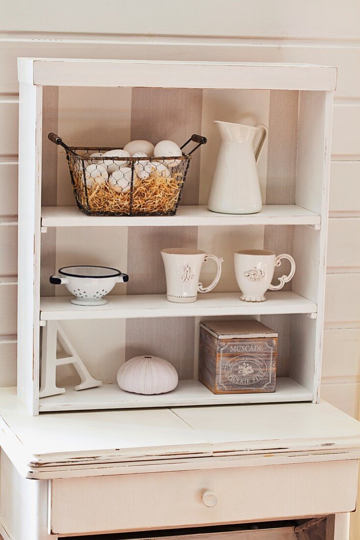 White crockery in small shelving unit on shabby-chic console table against white wooden wall