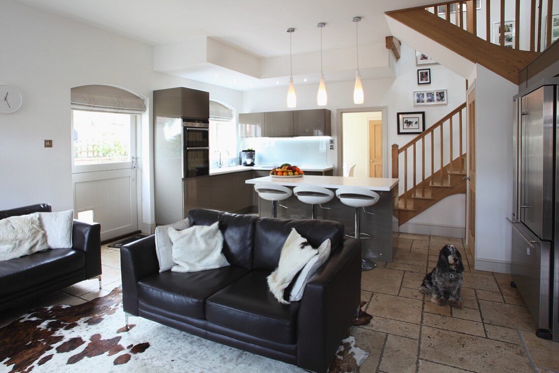 Leather sofas and cow-skin rug in open-plan interior with modern fitted kitchen and country house style staircase in background