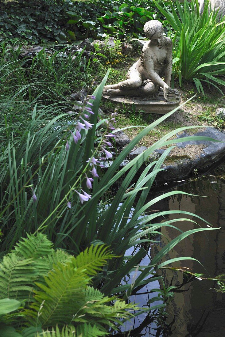 Sitzende Steinfigur zwischen Ziergräsern am Teich
