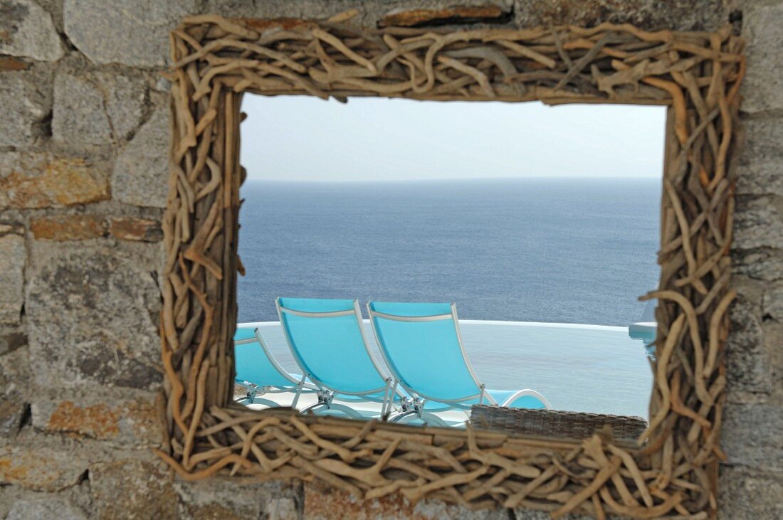 Spiegel mit Rahmen aus kleinen Zweigen und sich spiegelnde Liegestühle am Meer