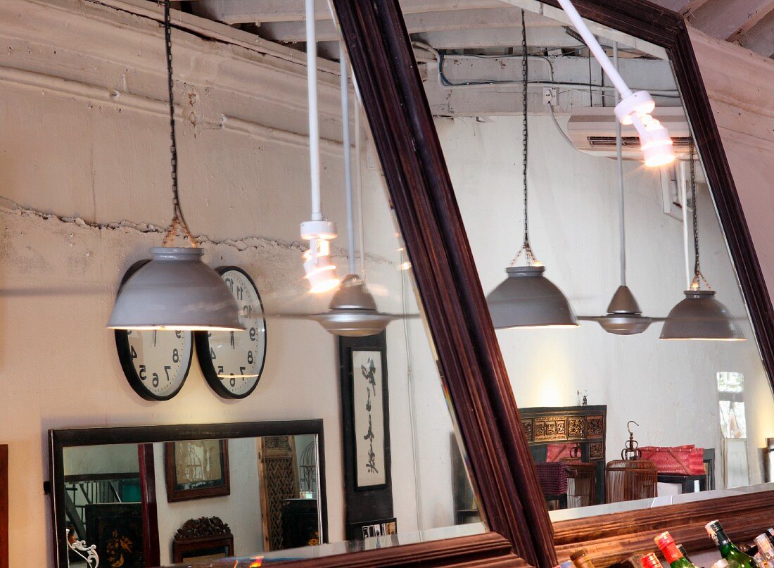 Hängeleuchten mit Metallschirm und Uhren an Wand im Innenraum eines Restaurants reflektiert in zwei großen Wandspiegeln